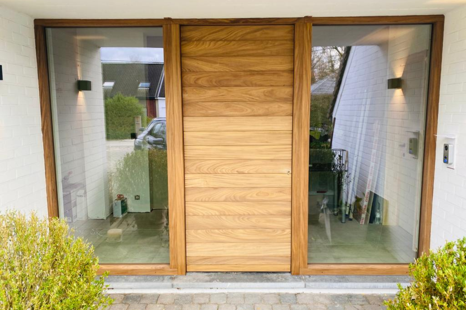 Moderne houten voordeur met verticale panelen, geflankeerd door twee grote glazen zijpanelen in een witte gevel.