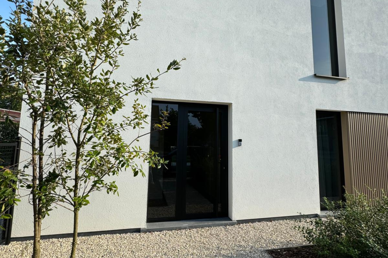 Moderne witte woning met strakke gevel, zwarte raamprofielen en een minimalistische voortuin met grindpad en jonge boom.