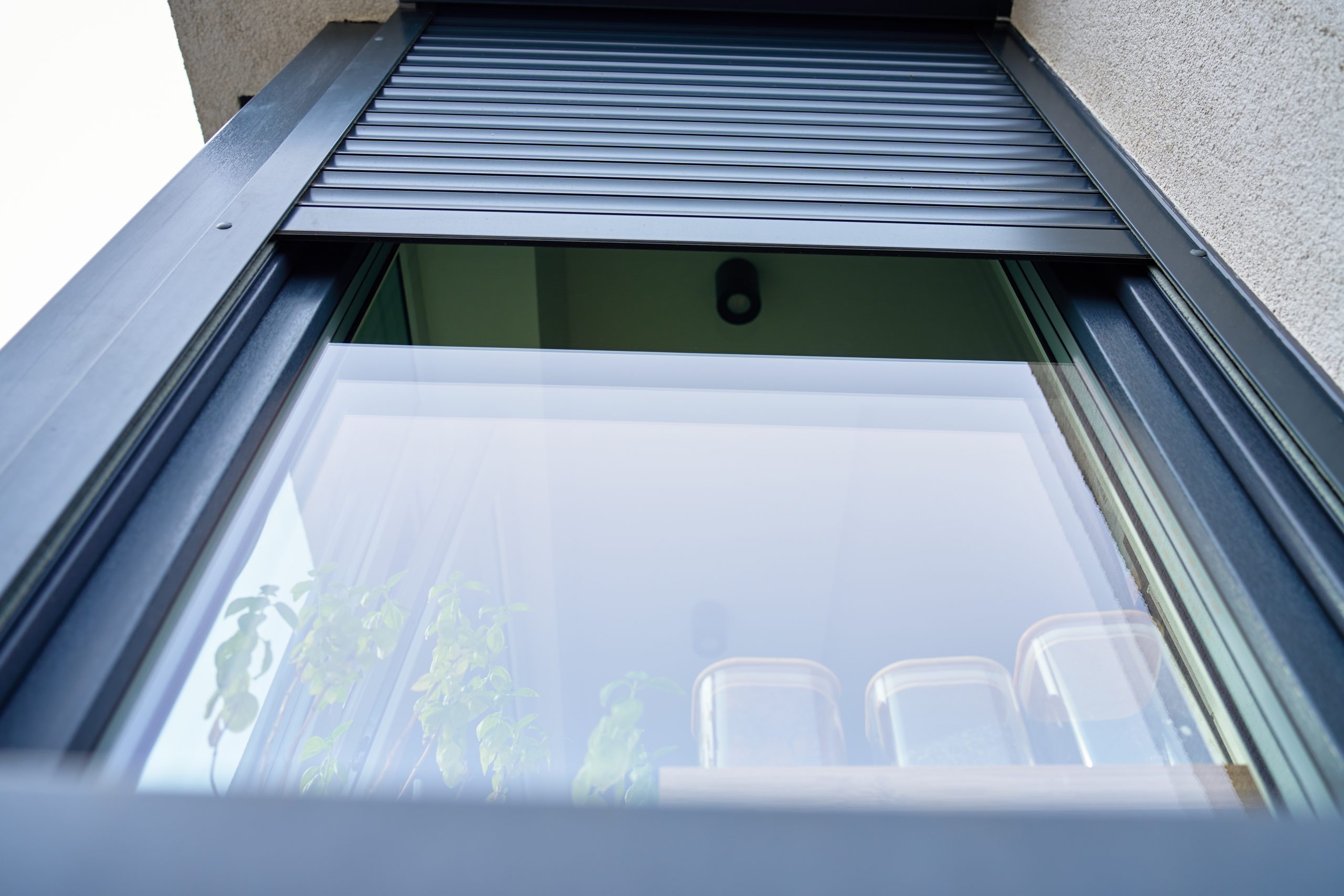 Antraciet aluminium opbouwrolluik half neergelaten op een raam, met zicht op de geleiders en de gevel.