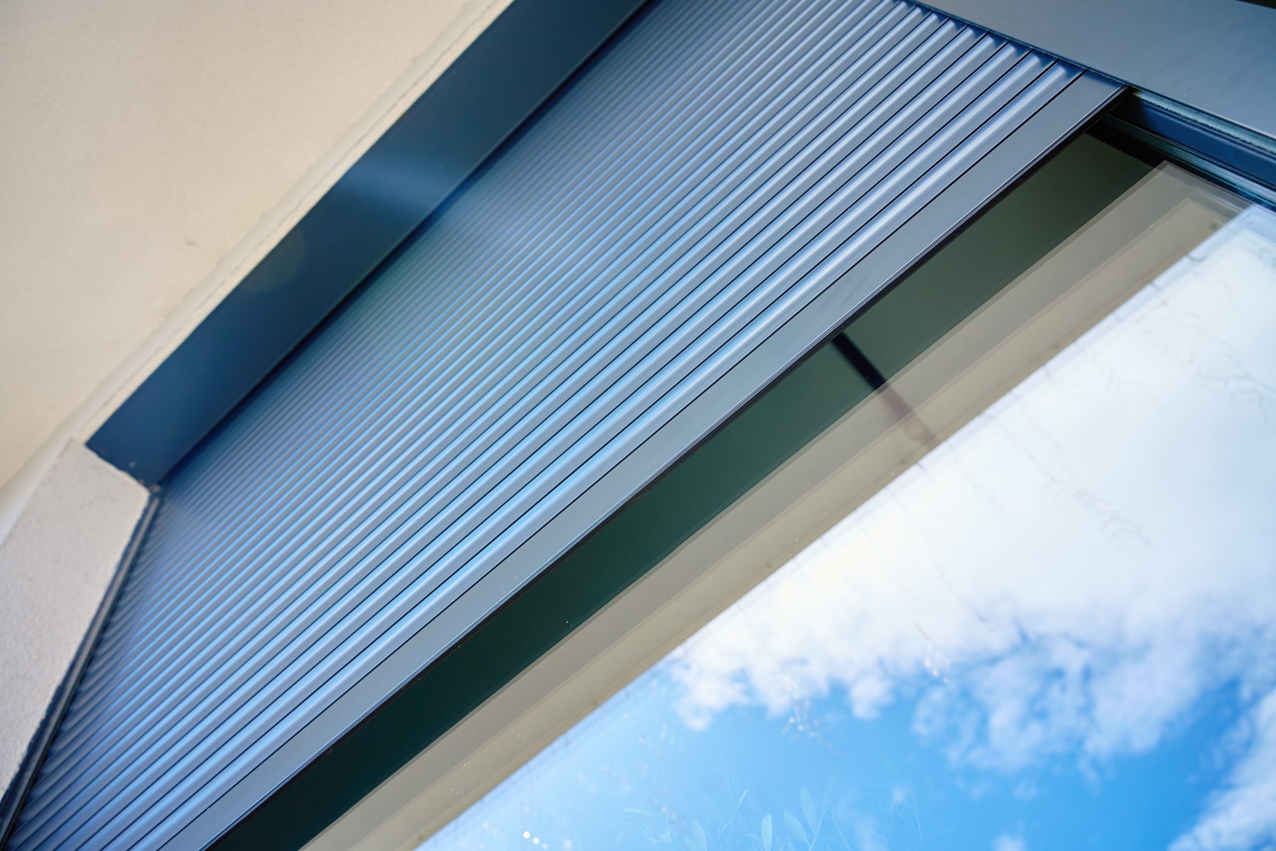 Aluminium rolluik half geopend op een modern raam, met zicht op de lamellen en de blauwe lucht eronder.