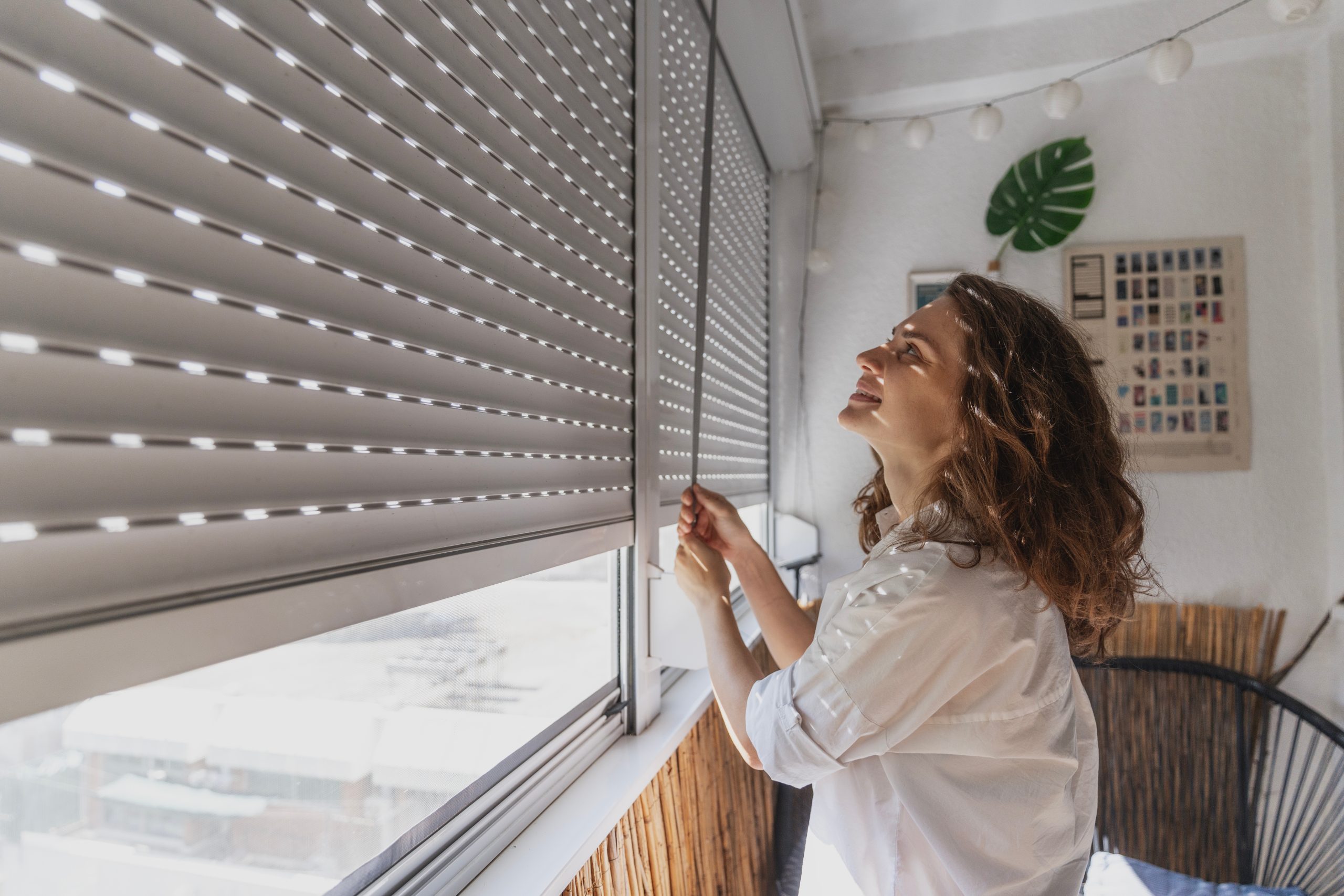 Vrouw die manueel een rolluik omhoogtrekt via het lint, van binnenuit een lichte woonruimte.