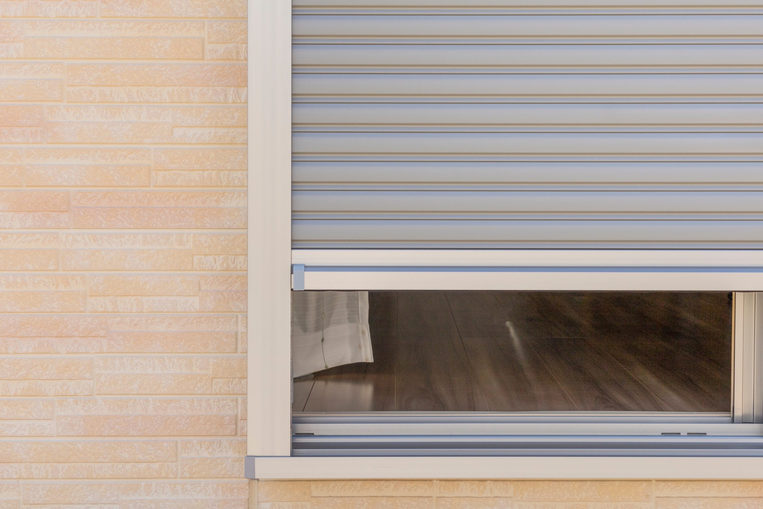 Wit PVC opbouwrolluik half neergelaten op een raam van een woning met lichte gevelsteen.
